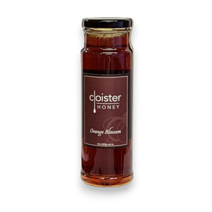 A tall, clear glass jar filled with dark amber honey labeled "Cloister Honey" with "Orange Blossom" beneath it on a brown label. The jar has a black metal lid and is photographed against a white background, highlighting the rich color and subtle citrus undertone of the natural Cloister Honey - Orange Blossom Honey 12oz.