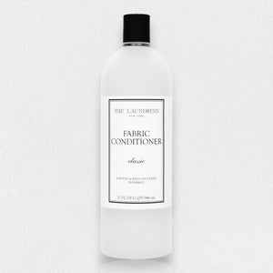 A white bottle of THE LAUNDRESS - FABRIC CONDITIONER 32OZ with a "Classic" scent label. This 32 fluid ounce (946 milliliters) fabric conditioner by THE LAUNDRESS promises to soften fabrics and reduce static. The bottle features a black cap, set against a light gray background.