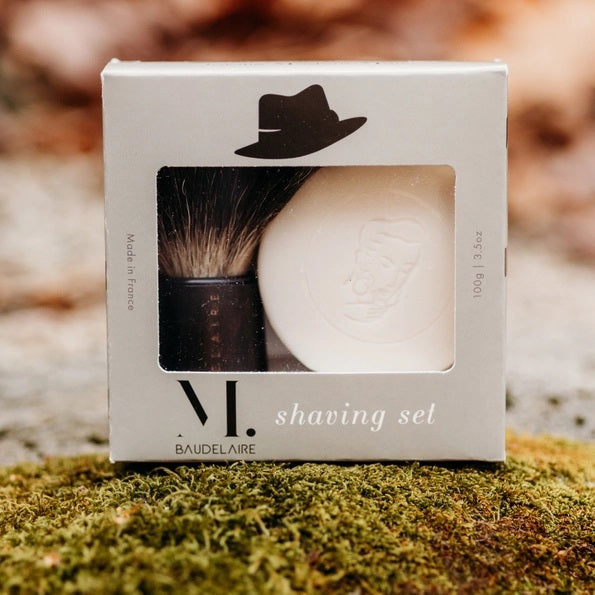 A hand holds the BAUDELAIRE - M.B. SHAVING SET bowl filled with foamy shaving soap, using the included badger bristle brush from BAUDELAIRE to whip up a rich lather.