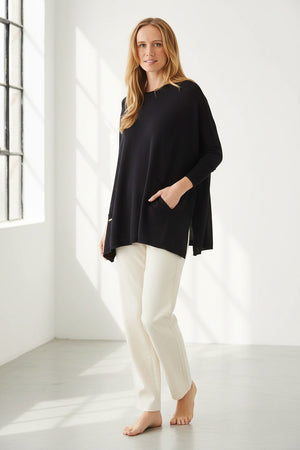 A smiling woman with long blonde hair wears the MERSEA Catalina Crewneck Sweater in black, featuring signature split sides, paired with light pants and standing against a plain white background.