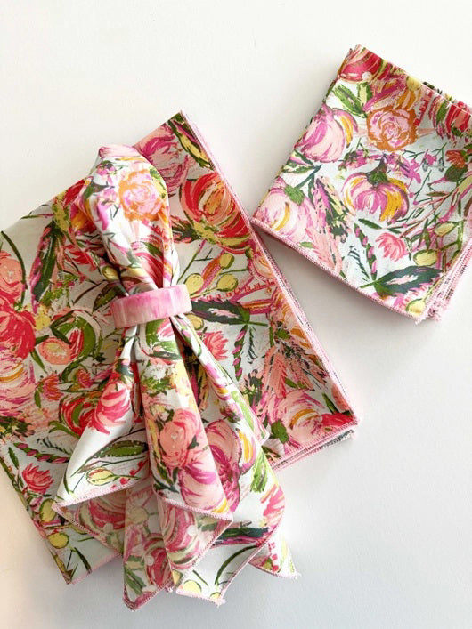 A set of FLORAL FROLIC DINNER NAPKINS by DOT AND ARMY, featuring a floral pattern on a white background. One napkin is tied neatly with a pink ribbon, another is folded, and the last one is partially visible. The super soft fabric showcases bright colors with pink, yellow, and green floral designs.