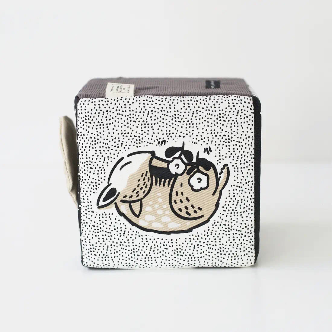 The WEE GALLERY - BABY ANIMALS SOFT BLOCK by WEE GALLERY is a fabric cube adorned with black and white designs that include adorable baby animals, such as a sleeping fox and its cub on one side. This sensory development toy features various textures, including polka dots and stripes, and includes two fabric loops on one edge to aid in tactile exploration.