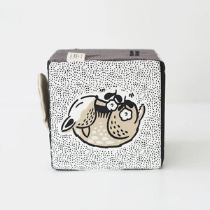 The WEE GALLERY - BABY ANIMALS SOFT BLOCK by WEE GALLERY is a fabric cube adorned with black and white designs that include adorable baby animals, such as a sleeping fox and its cub on one side. This sensory development toy features various textures, including polka dots and stripes, and includes two fabric loops on one edge to aid in tactile exploration.