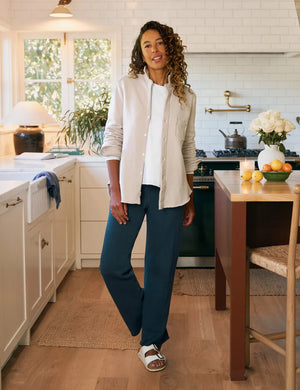 A woman with curly hair stands in a bright kitchen, wearing a white button-up and FRANK & EILEEN’s Catherine Long Sweatpant in Vintage Navy. The space has white cabinets, a stove, flowers in a vase, and a bowl of fruit on the counter.