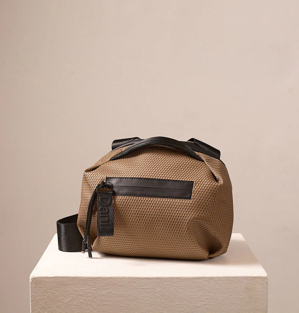 A person in a light beige suit carries the DANIELLA LEHAVI - BONNIE TRAVEL CROSSBODY, a large brown vegan bag with black zipper and strap, shown from shoulders to mid-thigh against a plain background.