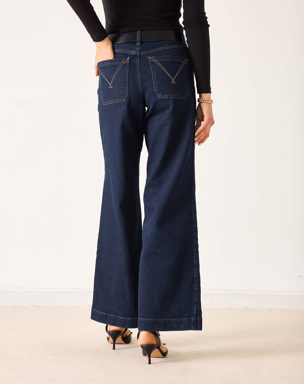 A person wearing MERSEA - SAILOR WIDE LEG JEANS IN RINSE by MERSEA, styled with a black top, black belt, and black heeled sandals, stands with their back to the camera against a plain light background.