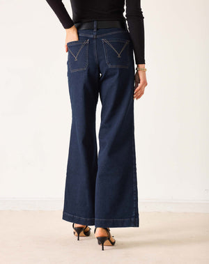 A person wearing MERSEA - SAILOR WIDE LEG JEANS IN RINSE by MERSEA, styled with a black top, black belt, and black heeled sandals, stands with their back to the camera against a plain light background.