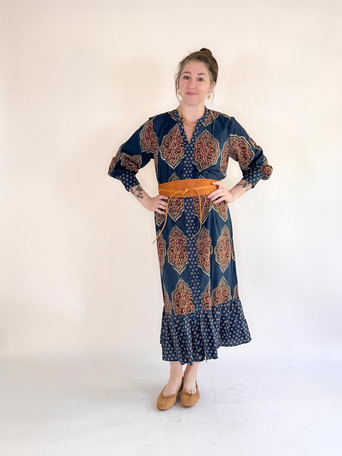 A woman stands against a plain background, smiling slightly with her hands on her hips, wearing the FARYLINA - GRACIE MAXI DRESS—a bohemian cotton dress with a ruffled hem, wide orange belt, and tan flats.