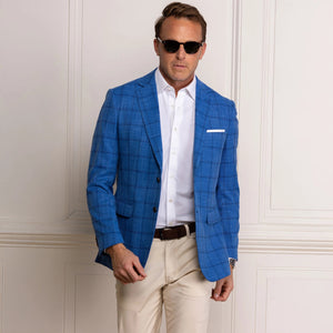 A man wearing sunglasses, the COLLARS AND CO MAVERICK TRAVEL BLAZER IN BLUE WINDOWPANE over a white t-shirt, along with light pants and a brown belt, stands smiling with one hand in his pocket against a white paneled wall.