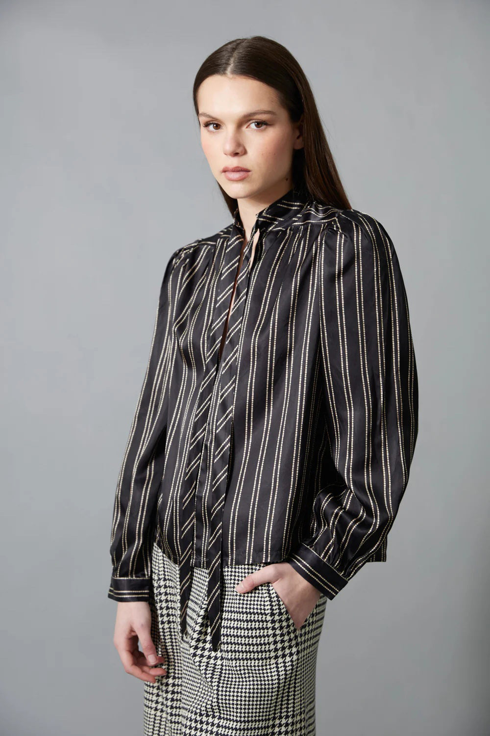 A woman with straight brown hair faces away in a HOPE FOR FLOWERS Tie Neck Blouse—black and white, vertically striped Tencel with puffed sleeves—paired with wide-leg plaid trousers. The plain gray background reflects sustainable style.