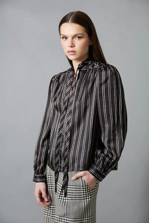 A woman with straight brown hair faces away in a HOPE FOR FLOWERS Tie Neck Blouse—black and white, vertically striped Tencel with puffed sleeves—paired with wide-leg plaid trousers. The plain gray background reflects sustainable style.