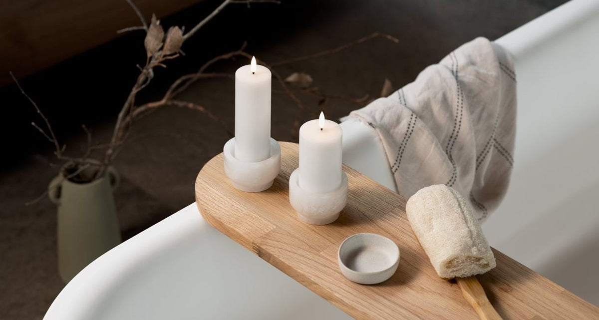 Three lit PIFFANY UYUNI Ivory Pillar Candles (2x3) are arranged on a wooden tray alongside ceramic decor and dishes atop a table. A small knife with a wooden handle rests nearby, while stacked bowls and a softly blurred sofa complete the minimal, cozy scene.