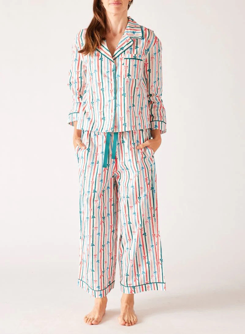 A woman stands barefoot in the MERSEA - OVER THE MOON PAJAMA SET, a 100% cotton ensemble by MERSEA with vibrant red, teal, and white vertical stripes and arrow patterns; she has her hands in her pockets and long hair over one shoulder.