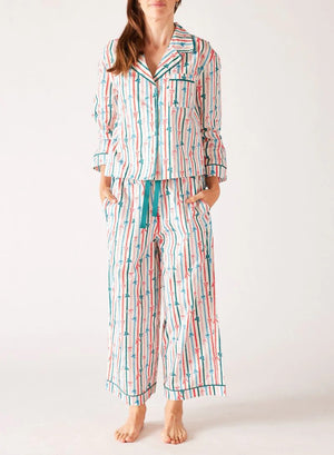 A woman stands barefoot in the MERSEA - OVER THE MOON PAJAMA SET, a 100% cotton ensemble by MERSEA with vibrant red, teal, and white vertical stripes and arrow patterns; she has her hands in her pockets and long hair over one shoulder.