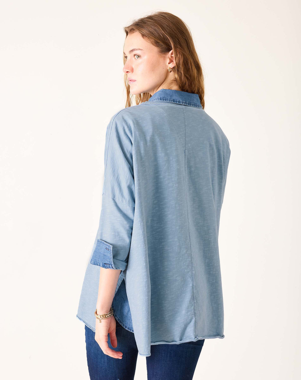A woman with wavy light brown hair wears the MERSEA - Catalina Slub Tee in Shore Blue Stitches with rolled-up sleeves over denim jeans, looking to her left against a plain, light background.