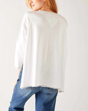 A woman with wavy brown hair models the MERSEA - CATALINA SLUB TEE—an oversized, long-sleeved white top with thin gray stripes—by MERSEA. She pairs it with blue jeans and stands against a plain, light background, hands behind her back.
