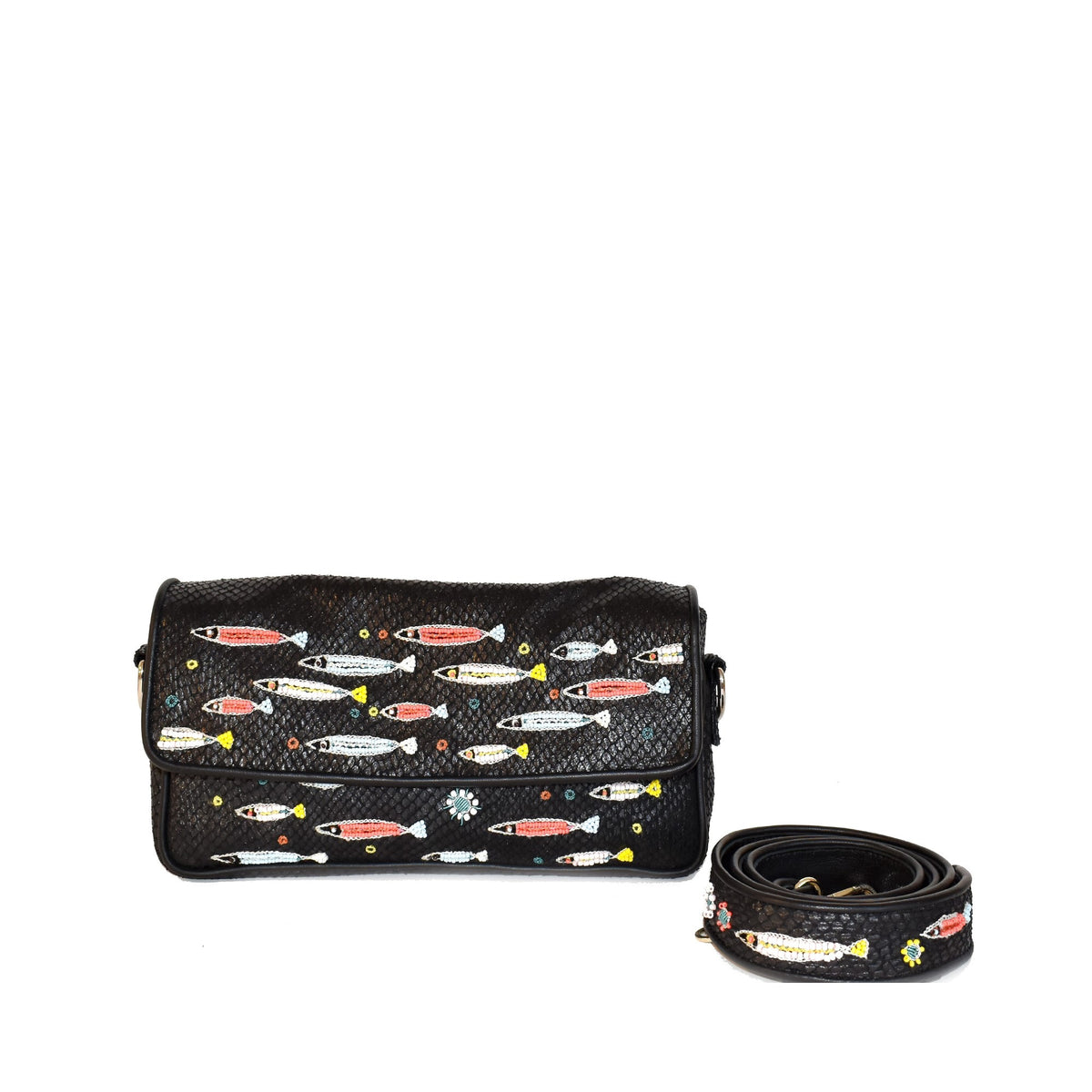 A woman with curly hair lies on a textured surface, wearing a light pink dress. She holds the MEHER KAKALIA Tavira Clutch Magma Black adorned with hand-embroidered fish motifs and wears black and white striped shoes. The soft lighting creates a relaxed, serene atmosphere.