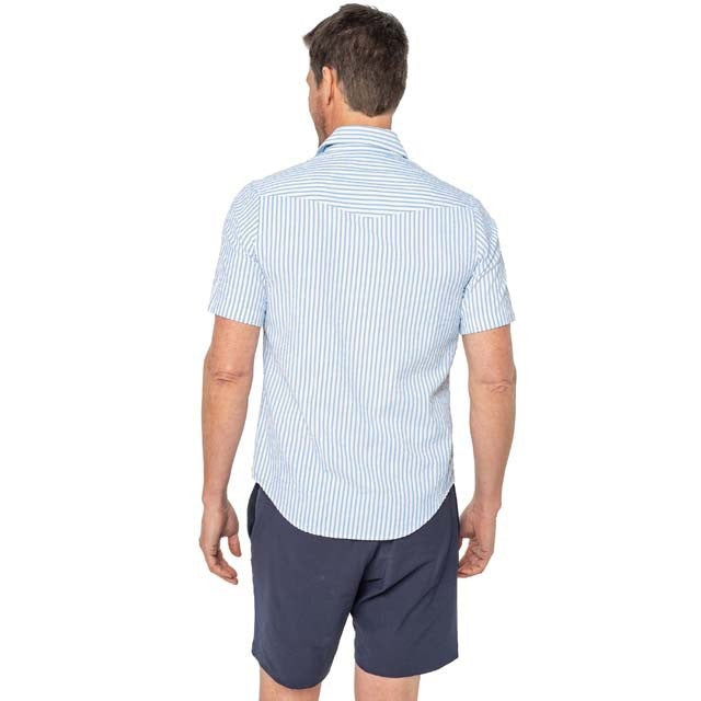 A man with a gray beard wears the CRIQUET Short Sleeve Performance Seersucker Pearl Snap Shirt in Navy while standing on a boat, with water, palm trees, and cloudy skies in the background.