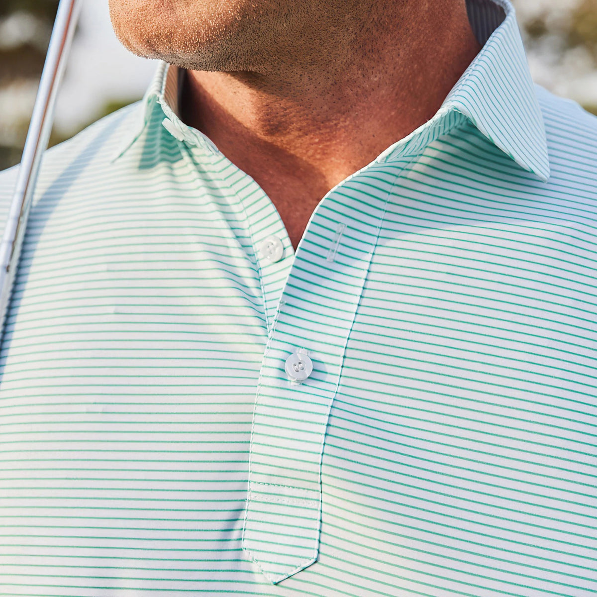 A man in sunglasses wears the COLLARS AND CO Featherweight Flex Polo in Augusta Green Stripe while standing outdoors on grass with trees behind him under a partly cloudy sky.