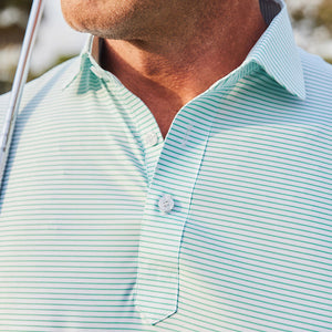 A man in sunglasses wears the COLLARS AND CO Featherweight Flex Polo in Augusta Green Stripe while standing outdoors on grass with trees behind him under a partly cloudy sky.