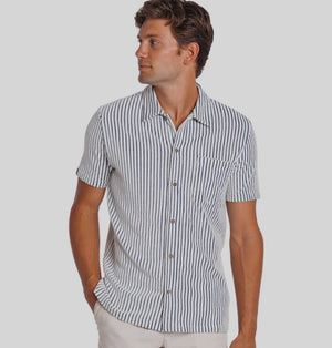 A man with curly hair and a short beard wears the CRIQUET Terrycloth Button Down Shirt in Striped Jacquard Natural, holding a drink in a koozie. Surfboards, sand, and trees create a relaxed summer vibe in the background.