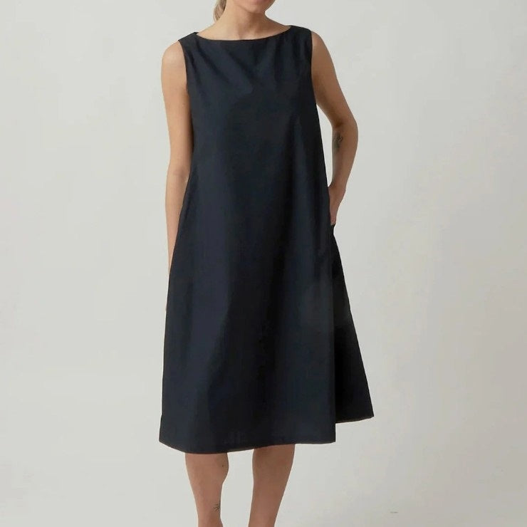 A person wearing the KATHARINA HOVMAN Sleeveless Dress in Heavy Taffeta—a loose-fitting, knee-length black dress—stands against a plain light background. Their arms are relaxed at their sides, and their head is out of frame.