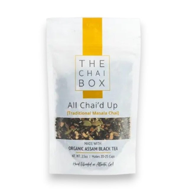 A white teapot pours tea into a glass cup beside a bag of THE CHAI BOX - ALL CHAI’D UP FULL BODY + BALANCE loose-leaf black tea from THE CHAI BOX, while a white vase with orange roses rests on a white surface near the window.