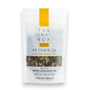 A white teapot pours tea into a glass cup beside a bag of THE CHAI BOX - ALL CHAI’D UP FULL BODY + BALANCE loose-leaf black tea from THE CHAI BOX, while a white vase with orange roses rests on a white surface near the window.