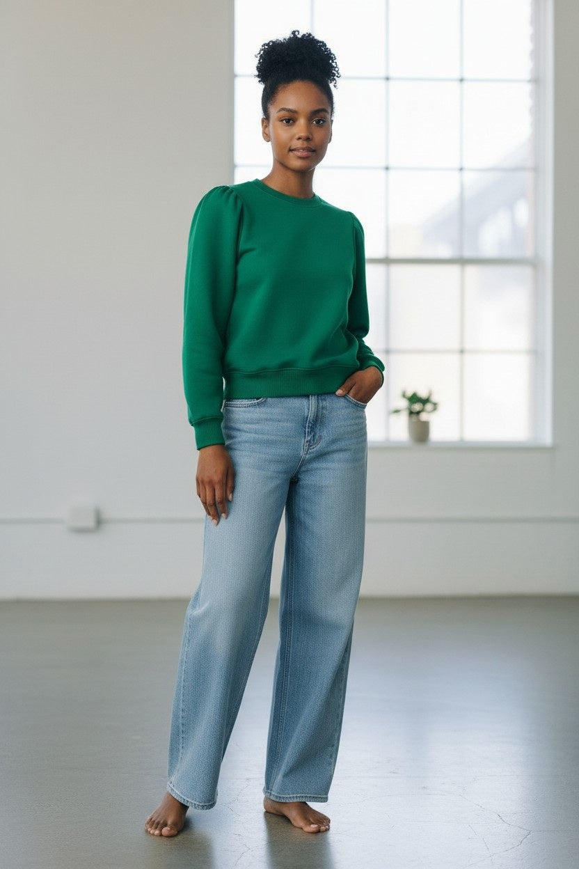 A woman with short gray hair stands against a plain wall, wearing the KMJ - COOPER SWEATSHIRT IN KELLY GREEN by KMJ, paired with light blue wide-leg jeans and white sneakers. She keeps her hands in her pockets and maintains a neutral expression.