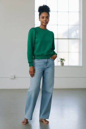 A woman with short gray hair stands against a plain wall, wearing the KMJ - COOPER SWEATSHIRT IN KELLY GREEN by KMJ, paired with light blue wide-leg jeans and white sneakers. She keeps her hands in her pockets and maintains a neutral expression.