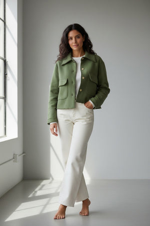 A woman with long, light brown hair stands in a doorway—one hand in her pocket—wearing the FRANK & EILEEN Rowan Cropped Sweatshirt Utility Jacket in Triple Fleece Nori paired with white pants. The background features a stable area and horse equipment.