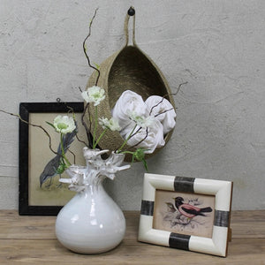 The HOMART CERAMIC VASE WITH BRANCH AND BIRD features white flowers and branches, set on a wooden surface with framed bird art and a woven basket against a light gray textured wall for a cozy touch.