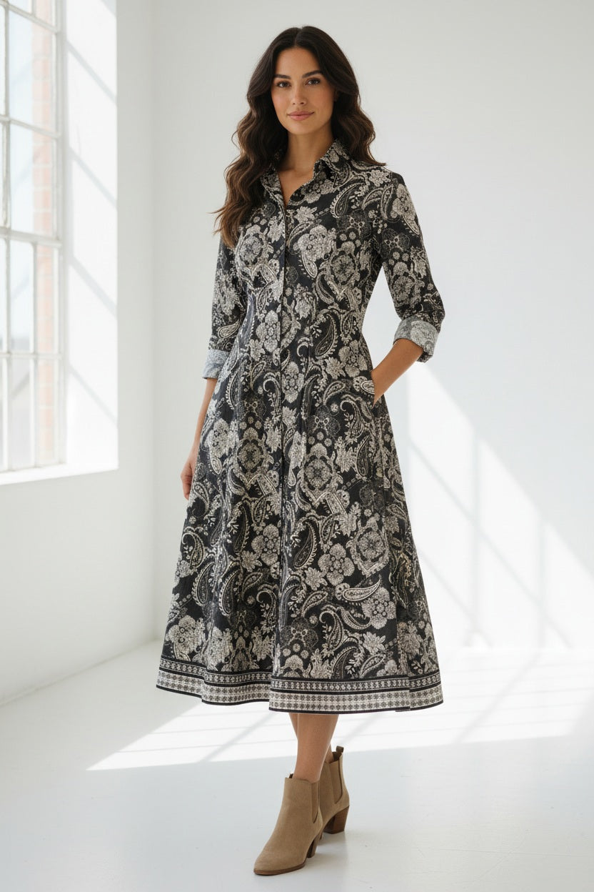 A woman stands wearing the HINSON WU - MARTINA 2/4 SLEEVE PAISLEY PRINT DRESS. This refined cotton dress by HINSON WU features a black background with a white floral and paisley print, a collared neckline, mid-calf length, flared skirt, side pockets, and elegant 2/4 sleeves.