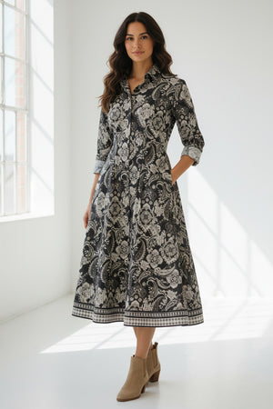 A woman stands wearing the HINSON WU - MARTINA 2/4 SLEEVE PAISLEY PRINT DRESS. This refined cotton dress by HINSON WU features a black background with a white floral and paisley print, a collared neckline, mid-calf length, flared skirt, side pockets, and elegant 2/4 sleeves.