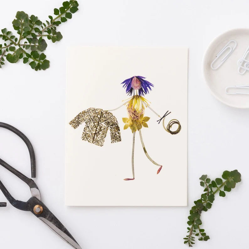 The PETAL PEOPLE PRESS - KNIT AND PURL GREETING CARD features a whimsical figure made from flower petals and leaves, with blue petal hair, a leafy jacket, and swirls of grass on a plain white background.