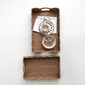 Two rectangular woven baskets from Creative Co-op are placed vertically on a white surface. The top basket, a BRAIDED BANKUAN TRAY WITH HANDLE (SMALL), contains a cup and saucer, a bowl with cereal, and some papers. The bottom basket is empty and has a slightly scalloped edge.