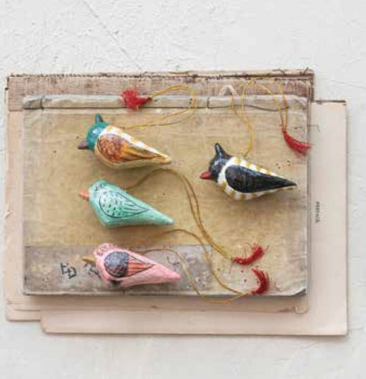 Four CREATIVE COOP PAPER MACHE BIRD ORNAMENTS in green, pink, brown, and black-and-white, with strings and red tassels, are shown on vintage papers against a light wall. Each ornament is hand painted and colorful.