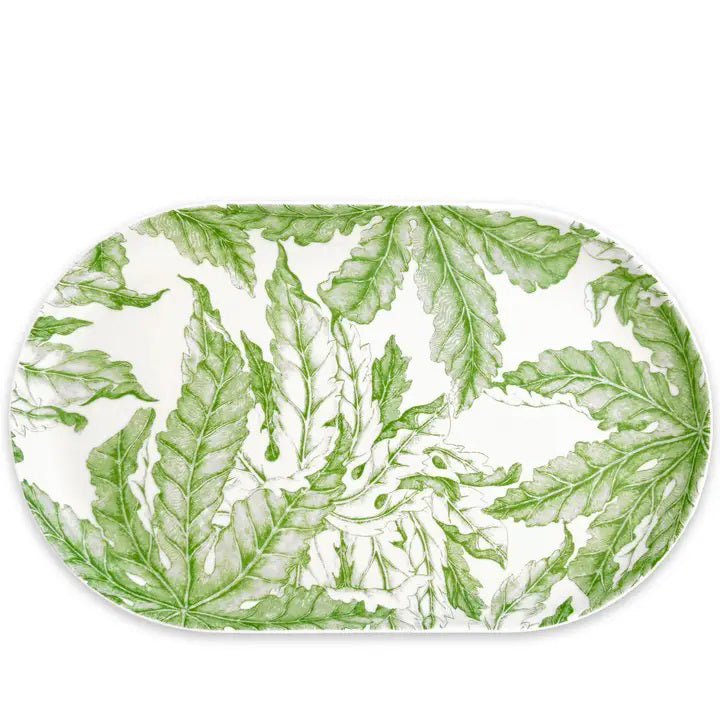 The CASKATA FREYA Coupe Oval Platter rests on a rustic green table amid carrots, grapes, greens, and white flowers. This porcelain piece features floral designs and is both dishwasher and microwave safe for easy care.