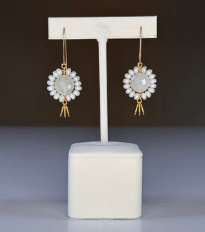 The GOLDISH - Floral Gemstone Earrings in Moonstone and White Pearl feature a circular, faceted moonstone center surrounded by white pearls, with three gold danglers, all set in 14K gold fill. Displayed on a white stand against a dark background.