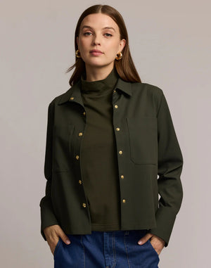 A woman with long brown hair models the HINSON WU - ASTRID LONG SLEEVE TRAVELER'S TWILL SHIRT JACKET in dark green over an olive top and blue jeans, showcasing versatile HINSON WU travel style with gold hoop earrings against a neutral background.