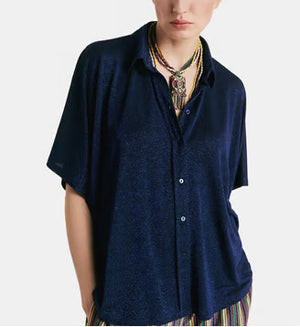 A person in a MOMONI - BROOKLYN blue shirt with a classic collar, paired with colorful beaded necklaces and tucked into striped pants. The loose-fitting, short-sleeve button-up stands out against the plain light background.