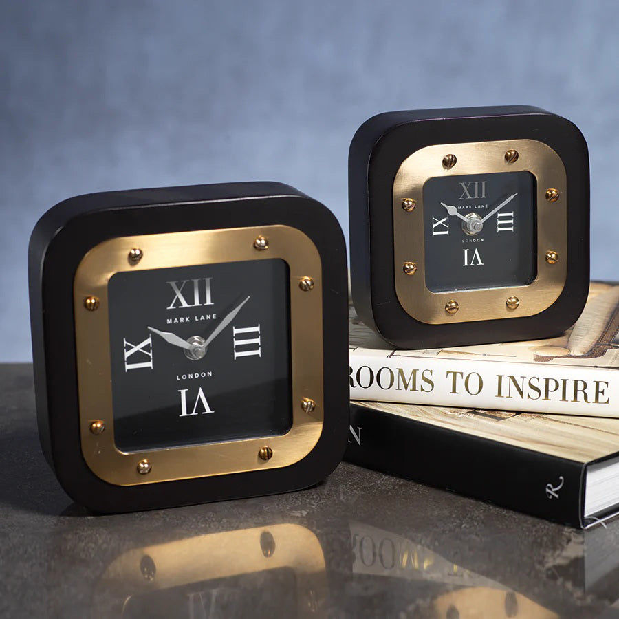 Two MARK LANE TABLE CLOCK - LARGE by ZODAX, square in shape with black and gold iron finish and Roman numerals, are displayed—one on a table, the other atop "Rooms to Inspire" books. The clocks show different times.