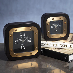 Two MARK LANE TABLE CLOCK - LARGE by ZODAX, square in shape with black and gold iron finish and Roman numerals, are displayed—one on a table, the other atop "Rooms to Inspire" books. The clocks show different times.