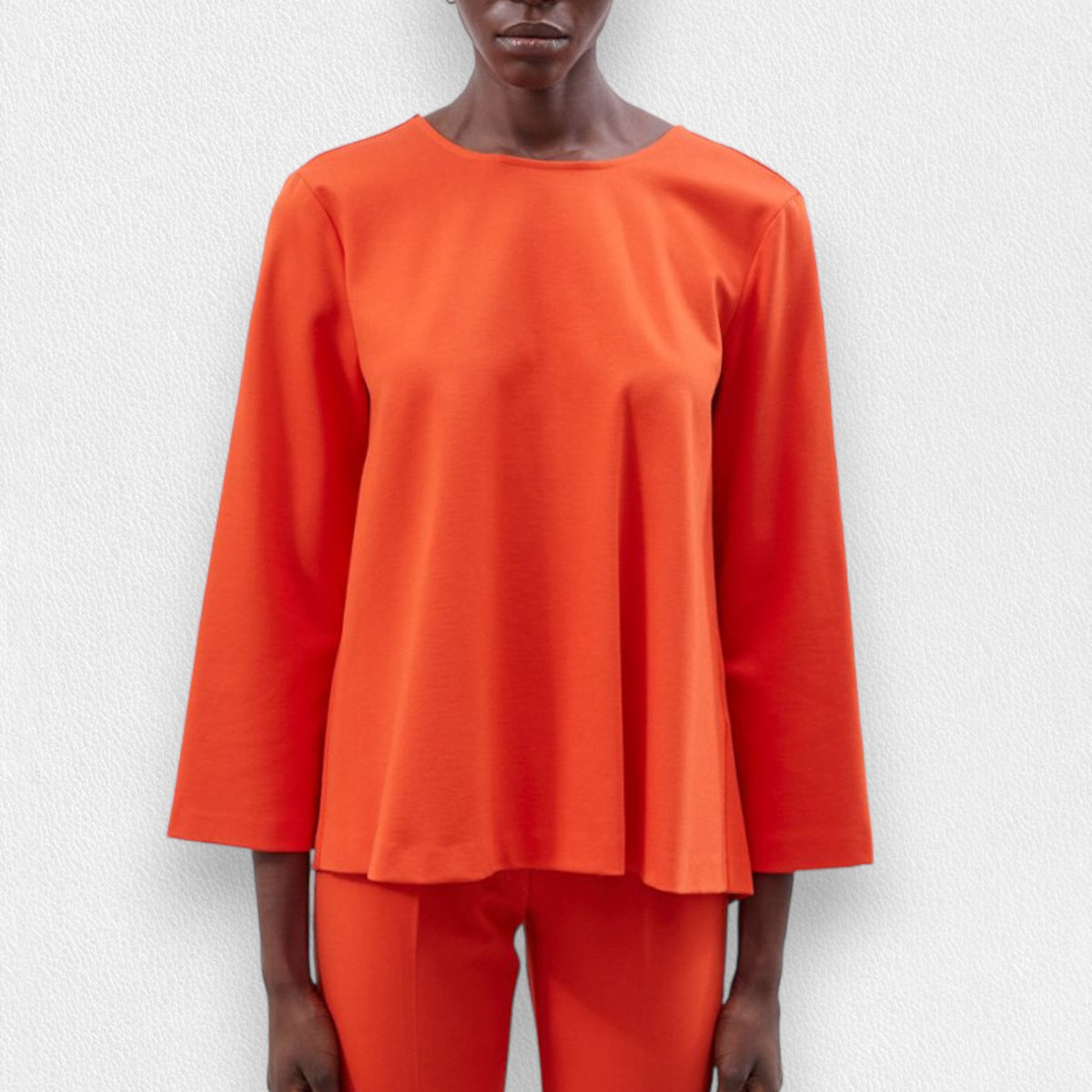 A person is wearing the MEIMEIJ - TOP WITH KNOT BACK DETAIL in orange, featuring wide sleeves and a stylish knot back design, paired with matching relaxed-fit orange pants. The outfit appears loose and comfortable, perfect for a casual look. The background is a plain, light-colored texture.