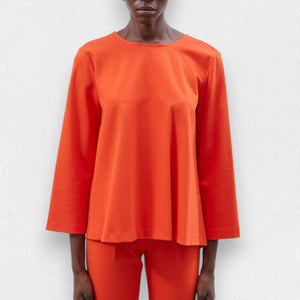 A person is wearing the MEIMEIJ - TOP WITH KNOT BACK DETAIL in orange, featuring wide sleeves and a stylish knot back design, paired with matching relaxed-fit orange pants. The outfit appears loose and comfortable, perfect for a casual look. The background is a plain, light-colored texture.