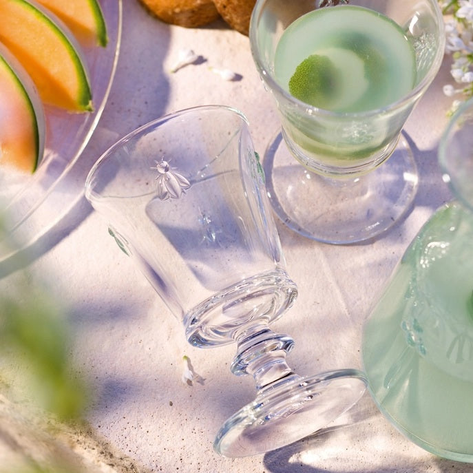 The BEE WATER GLASS by LA ROCHERE is a clear glass goblet with a thick stem and embossed bee pattern around the bowl, inspired by classic Napoleonic styles.