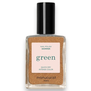 A hand adorned with metallic gold vegan nail polish holds a bottle of MANUCURIST - GREEN™ NAIL POLISH IN SHIMMER by MANUCURIST INC, set against a textured, light-filled background.
