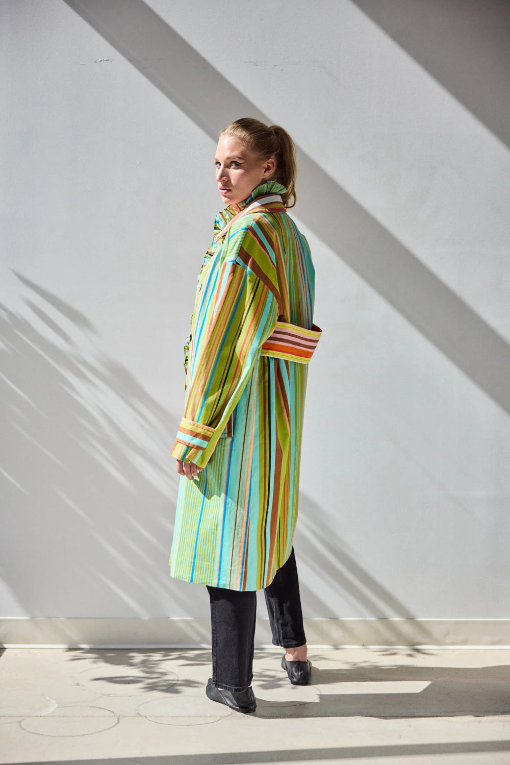 A person stands against a white wall wearing the HOPE FOR FLOWERS - COMBO STRIPES COAT by HOPE FOR FLOWERS, styled over a patterned shirt and black pants. Sunlight casts leaf-like shadows on the wall and floor behind them.