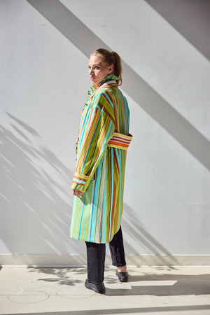 A person stands against a white wall wearing the HOPE FOR FLOWERS - COMBO STRIPES COAT by HOPE FOR FLOWERS, styled over a patterned shirt and black pants. Sunlight casts leaf-like shadows on the wall and floor behind them.