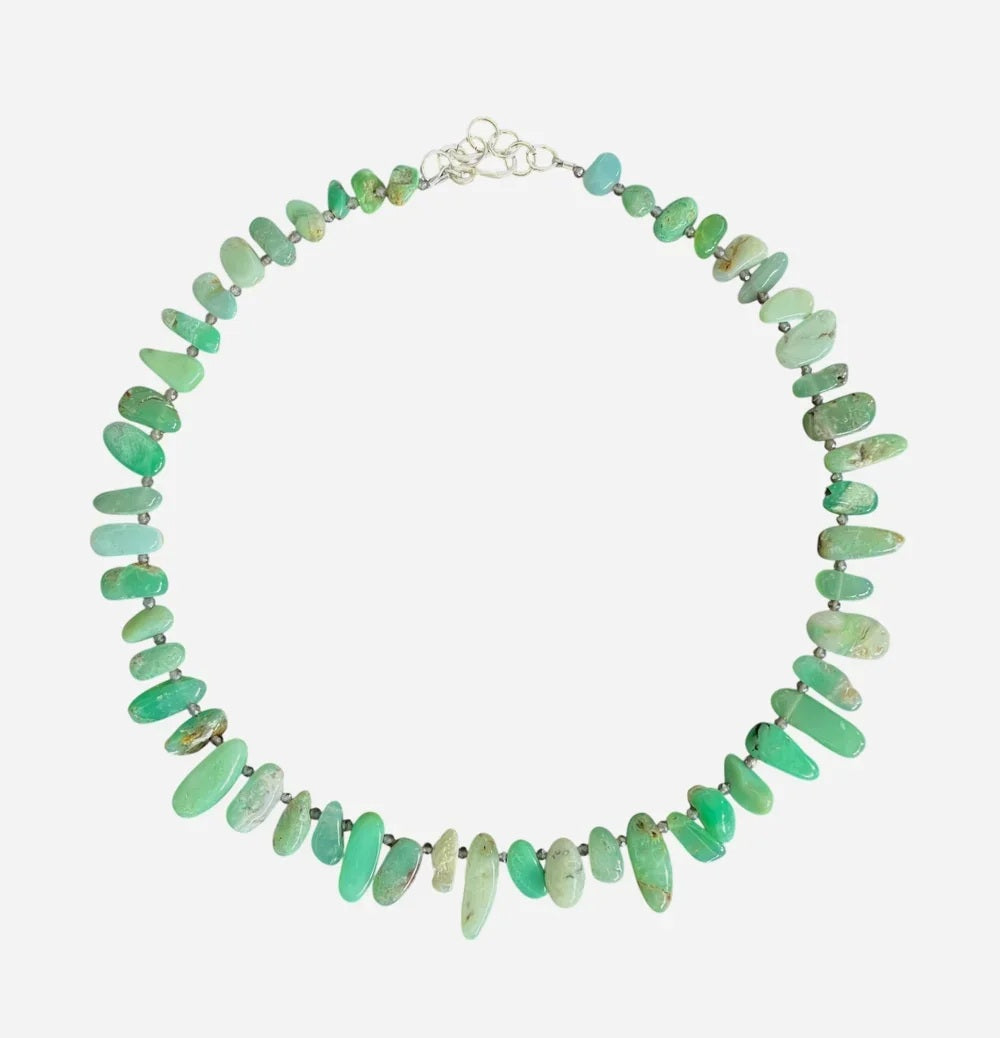 The ANN LIGHTFOOT Ras du Cou Necklace of Chrysoprase Pebbles features irregularly shaped green chrysoprase stones and a sterling silver clasp, elegantly displayed in a circular arrangement on a white background.